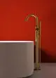 Freistehender Wasserhahn in gebürstetem Goldfinish von Ideal Standard, platziert neben einer runden weißen Badewanne vor einer roten Wand.